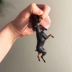 May include: A black and tan Doberman Pinscher keychain. The dog is standing on its hind legs, with its front paws raised. The keychain has a black chain and a gold-colored clasp.