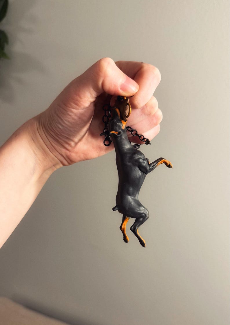 May include: A black and tan Doberman Pinscher keychain. The dog is standing on its hind legs, with its front paws raised. The keychain has a black chain and a gold-colored clasp.