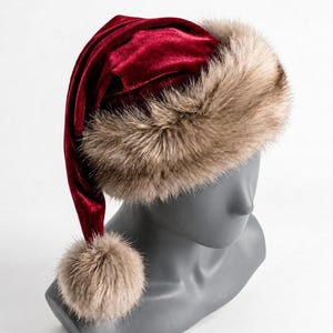 Puede incluir: Un gorro de terciopelo rojo intenso con un borde ancho de piel sint&eacute;tica beige esponjosa en la base y un pomp&oacute;n de piel sint&eacute;tica beige a juego en la punta. El gorro se muestra sobre una cabeza de maniqu&iacute; gris, resaltando su textura.