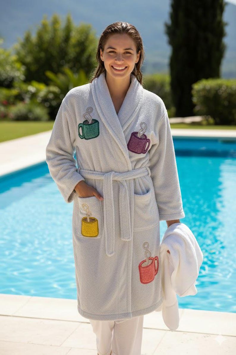 May include: A light grey terry cloth dressing gown featuring embroidered coffee cup designs in green, purple, yellow, and red. The gown has a shawl collar, pockets, and a matching belt. A white towel is held.