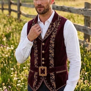 May include: A deep burgundy velvet vest featuring intricate gold floral embroidery along the lapels and front panels. The garment includes a row of round gold buttons and a matching gold buckle belt at the waist, worn over a white long-sleeve shirt.