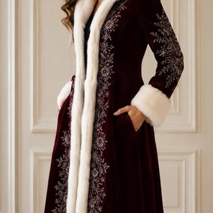 May include: A floor-length, deep burgundy velvet coat featuring a white faux fur trim along the hood, front opening, and sleeve cuffs. The coat is adorned with intricate silver and red floral embroidery along the front panels and sleeves.