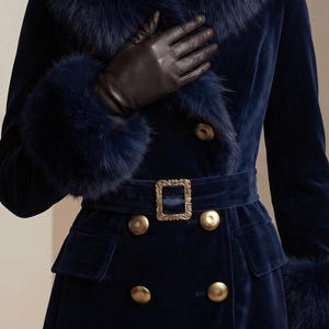 May include: A navy blue velvet coat featuring a large faux fur collar and matching fur cuffs. The coat is double-breasted with gold-toned buttons and a belted waist secured by a rectangular gold buckle. Dark brown leather gloves are worn.