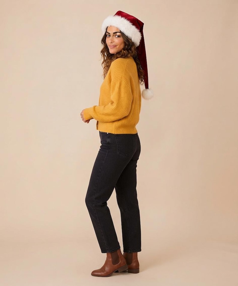 May include: A mustard yellow ribbed knit cardigan, black denim jeans, brown leather ankle boots, and a deep red velvet Santa hat with a white faux fur trim and a white pom-pom at the end of the long, pointed cap.
