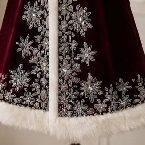 May include: A deep burgundy velvet cape featuring intricate silver thread embroidery in snowflake and floral patterns, accented with clear crystals. The garment is finished with a thick, white faux fur trim along the front edge and hemline.