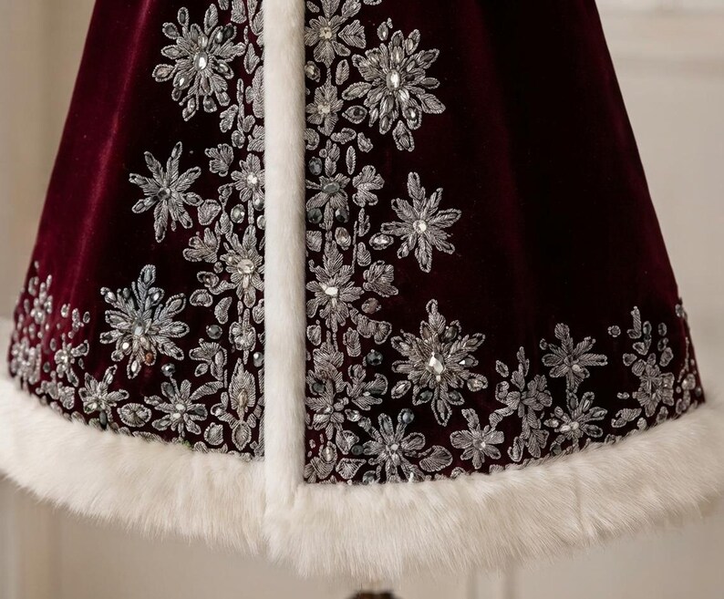 May include: A deep burgundy velvet cape featuring intricate silver thread embroidery in snowflake and floral patterns, accented with clear crystals. The garment is finished with a thick, white faux fur trim along the front edge and hemline.