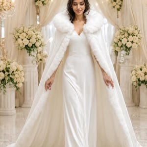 May include: A floor-length white gown paired with a matching white cape featuring a thick faux fur trim along the hood and front edges. The cape drapes fully to the floor, creating a long silhouette that complements the simple, sleeveless dress.