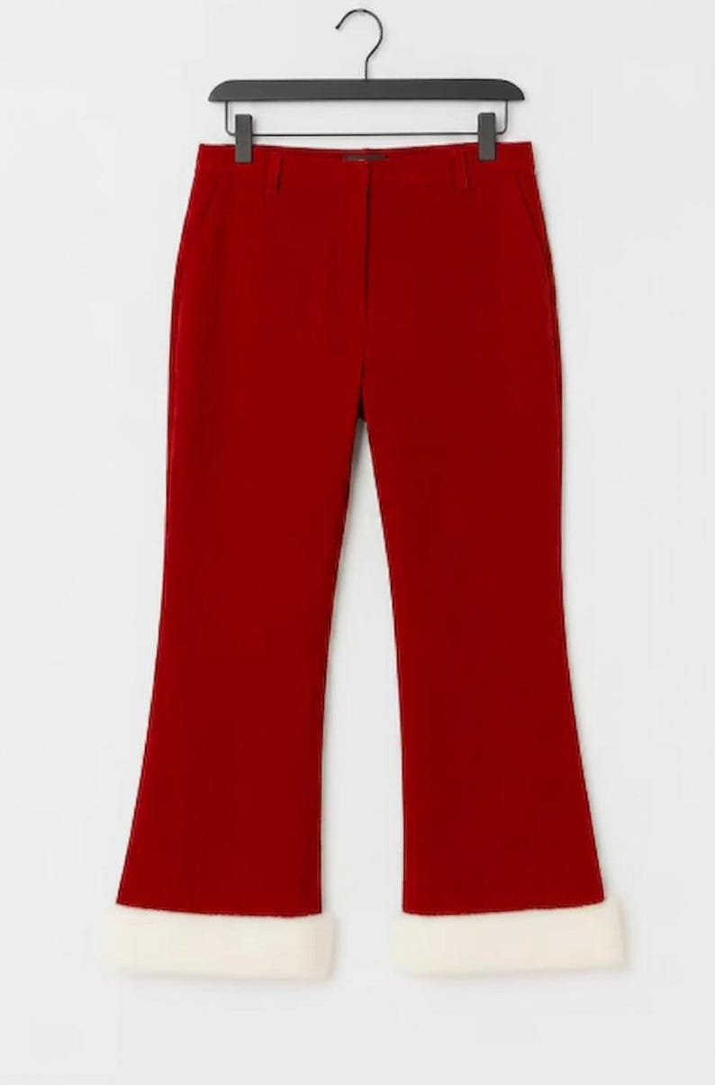 May include: A pair of solid red trousers featuring a straight-leg cut and a wide, plush white faux fur trim at the bottom of each leg. The trousers include a standard waistband with belt loops and a central zip fly, hanging from a black hanger.