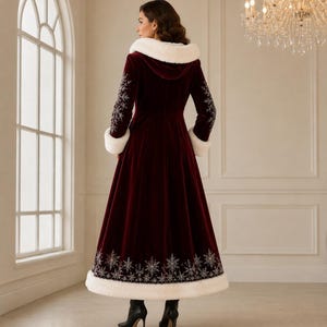 May include: A floor-length, deep burgundy velvet coat featuring a white faux fur hood trim and cuffs. The coat is adorned with intricate silver snowflake embroidery along the sleeves and the lower hemline, paired with black heeled ankle boots.
