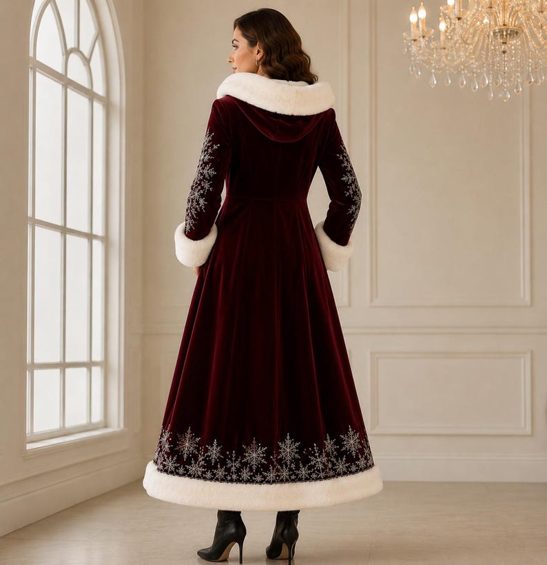 May include: A floor-length, deep burgundy velvet coat featuring a white faux fur hood trim and cuffs. The coat is adorned with intricate silver snowflake embroidery along the sleeves and the lower hemline, paired with black heeled ankle boots.