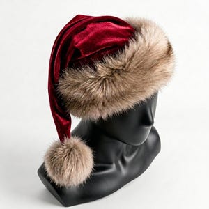 Puede incluir: Un gorro de terciopelo rojo intenso con una banda ancha y gruesa de piel sint&eacute;tica de color marr&oacute;n claro en la base. El gorro tiene una punta larga que cae hacia un lado, rematada con un pomp&oacute;n de piel sint&eacute;tica a juego.