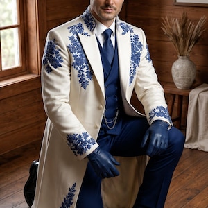 Puede incluir: Un abrigo largo de color crema con intrincados bordados florales azules en las solapas, los hombros y los pu&ntilde;os. El abrigo se lleva sobre un chaleco azul oscuro, una camisa blanca de cuello, una corbata azul, pantalones azul oscuro y guantes de cuero azul.