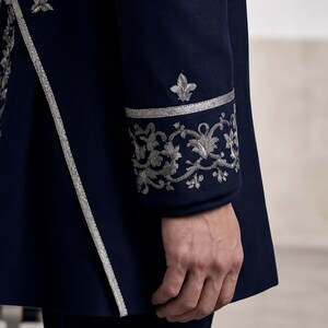 Puede incluir: Una chaqueta azul marino oscuro con intrincados bordados plateados en el pu&ntilde;o y a lo largo del borde frontal. El pu&ntilde;o muestra un patr&oacute;n floral detallado y un motivo de flor de lis, complementado por un ribete plateado en la abertura.