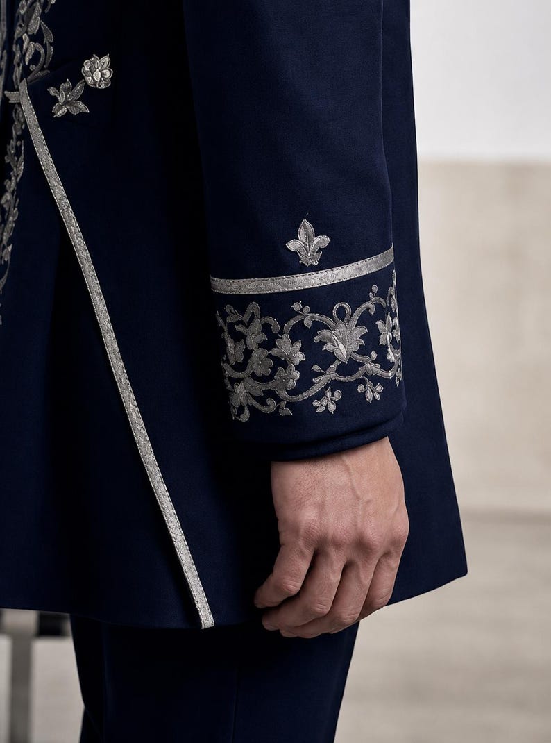 Puede incluir: Una chaqueta azul marino oscuro con intrincados bordados plateados en el pu&ntilde;o y a lo largo del borde frontal. El pu&ntilde;o muestra un patr&oacute;n floral detallado y un motivo de flor de lis, complementado por un ribete plateado en la abertura.