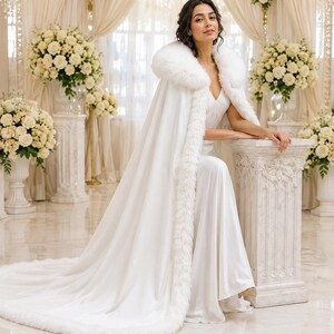 May include: A floor-length white cape featuring a thick, plush white faux fur trim along the hood and front edges. The cape is worn over a matching white sleeveless gown with a V-neckline, creating a cohesive, elegant, and layered ensemble.