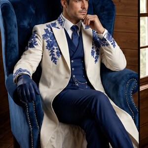 Puede incluir: Un abrigo largo de color crema con intrincados bordados florales azules en el cuello y los hombros, sobre un traje de tres piezas azul marino con corbata a juego, camisa blanca y guantes de cuero azul oscuro con cadena de bolsillo plateada.