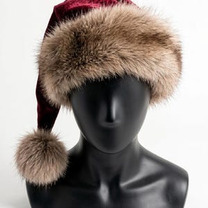 Puede incluir: Un gorro de terciopelo color burdeos intenso con un borde ancho de piel sint&eacute;tica de color marr&oacute;n claro y un pomp&oacute;n de piel sint&eacute;tica a juego en la punta, exhibido sobre una cabeza de maniqu&iacute; negra lisa.