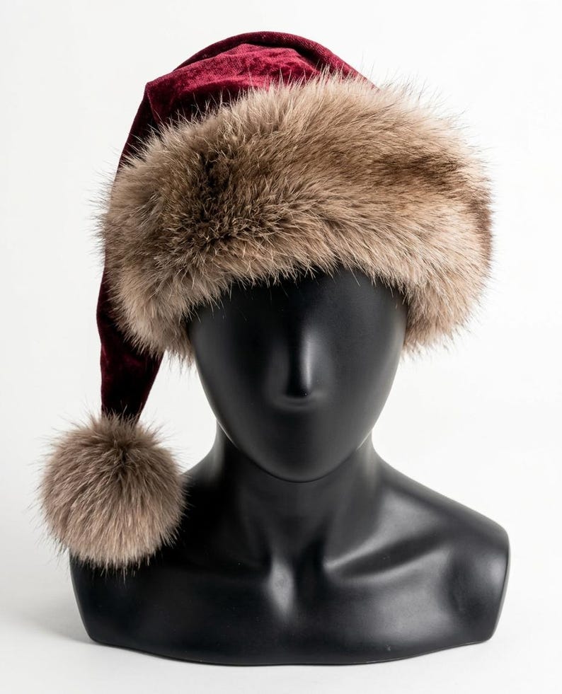 Puede incluir: Un gorro de terciopelo color burdeos intenso con un borde ancho de piel sint&eacute;tica de color marr&oacute;n claro y un pomp&oacute;n de piel sint&eacute;tica a juego en la punta, exhibido sobre una cabeza de maniqu&iacute; negra lisa.