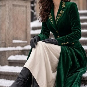 May include: A long-sleeved, dark green velvet coat featuring gold-toned buttons and intricate gold embroidery on the lapels. The coat is worn over a long, cream-coloured skirt, paired with black leather gloves and black leather heeled boots.