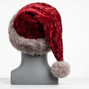 Puede incluir: Un gorro de terciopelo arrugado de color rojo intenso con un borde grueso y esponjoso de piel sint&eacute;tica gris. El gorro presenta una parte superior larga y puntiaguda que cuelga hacia un lado, terminada con un pomp&oacute;n de piel gris.
