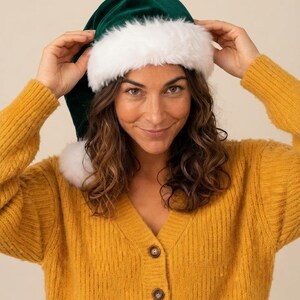 May include: A dark green velvet hat with a thick white faux fur trim around the base and a white faux fur pom-pom at the tip. The hat features a soft, plush texture and a classic conical shape, designed to be worn on the head.