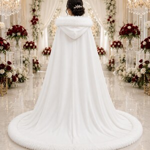 May include: A floor-length white hooded cloak featuring a thick faux fur trim along the hood and the entire circular hemline. The garment is made of a smooth, solid white fabric that drapes down from the shoulders to the floor in a full sweep.