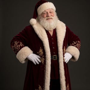 May include: A Santa Claus figure wearing a deep burgundy velvet coat with thick cream-colored faux fur trim and gold embroidery. The outfit includes a matching burgundy hat with a white pom-pom, a dark belt with a gold buckle, and white gloves.