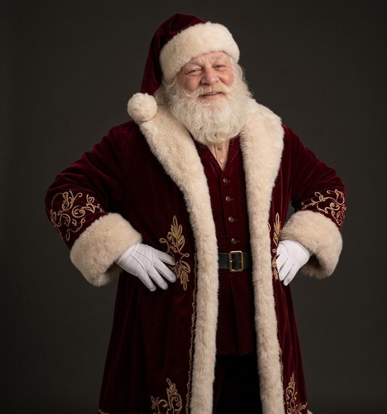May include: A Santa Claus figure wearing a deep burgundy velvet coat with thick cream-colored faux fur trim and gold embroidery. The outfit includes a matching burgundy hat with a white pom-pom, a dark belt with a gold buckle, and white gloves.
