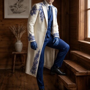 Puede incluir: Un abrigo largo de color crema con intrincados bordados florales azules en las solapas, pu&ntilde;os y dobladillo, usado sobre un chaleco, camisa y pantalones de color azul s&oacute;lido, junto con guantes de cuero azul y zapatos de vestir negros.