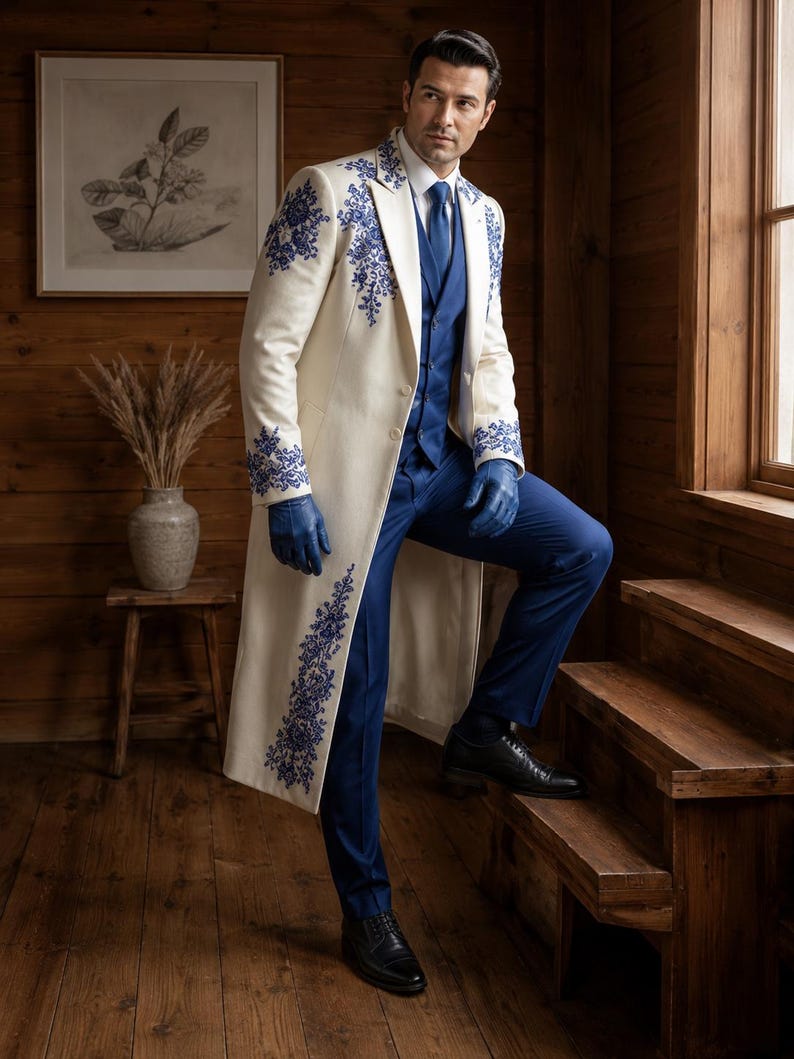 Puede incluir: Un abrigo largo de color crema con intrincados bordados florales azules en las solapas, pu&ntilde;os y dobladillo, usado sobre un chaleco, camisa y pantalones de color azul s&oacute;lido, junto con guantes de cuero azul y zapatos de vestir negros.