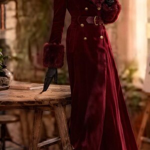 May include: A long, deep red velvet coat featuring a wide faux fur collar and matching faux fur cuffs. The coat is double-breasted with gold-toned buttons, a matching fabric belt with a gold buckle, and is paired with black leather gloves and boots.