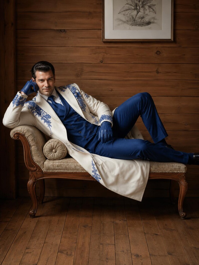 Puede incluir: Un abrigo largo de color crema con bordados florales azules en los hombros y pu&ntilde;os, sobre un chaleco azul, camisa blanca de cuello, corbata azul y pantalones azules. El conjunto se completa con guantes de cuero azul y zapatos de vestir oscuros.