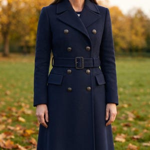 May include: A navy blue double-breasted coat featuring a structured collar, long sleeves, and a matching fabric belt with a rectangular buckle. The coat is adorned with two vertical rows of metallic buttons and includes two flap pockets at the waist.