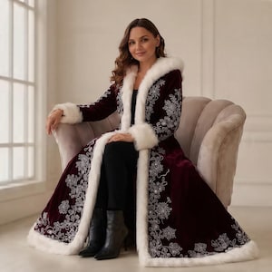 May include: A long, deep burgundy velvet coat featuring white faux fur trim along the hood, front opening, and cuffs. The coat is decorated with intricate silver floral embroidery patterns along the front panels and sleeves, worn over black trousers.