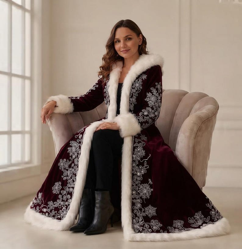 May include: A long, deep burgundy velvet coat featuring white faux fur trim along the hood, front opening, and cuffs. The coat is decorated with intricate silver floral embroidery patterns along the front panels and sleeves, worn over black trousers.