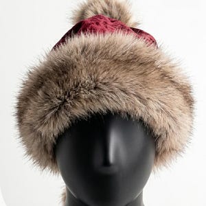 Puede incluir: Un gorro de terciopelo color burdeos intenso con una banda ancha y gruesa de piel sint&eacute;tica marr&oacute;n claro en la base y un pomp&oacute;n de piel sint&eacute;tica a juego en la parte superior, exhibido sobre una cabeza de maniqu&iacute; negra lisa.