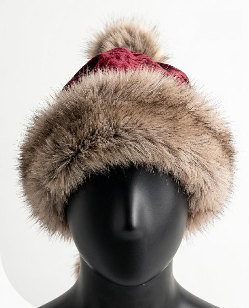 Puede incluir: Un gorro de terciopelo color burdeos intenso con una banda ancha y gruesa de piel sint&eacute;tica marr&oacute;n claro en la base y un pomp&oacute;n de piel sint&eacute;tica a juego en la parte superior, exhibido sobre una cabeza de maniqu&iacute; negra lisa.