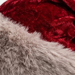 Puede incluir: Una vista de cerca de un gorro festivo con una corona de tela de terciopelo arrugado de color rojo intenso y un borde grueso de piel sint&eacute;tica beige texturizada en la parte inferior. El material tiene un brillo suave y aspecto denso.