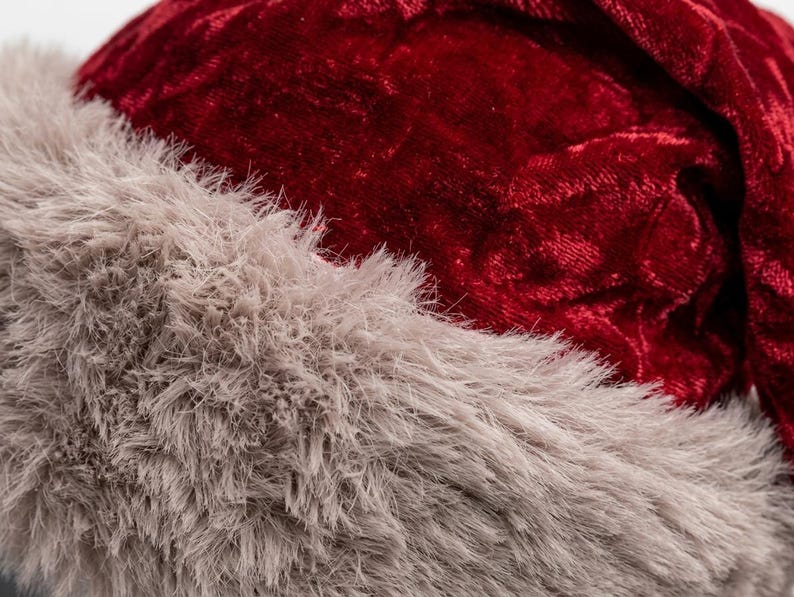 Puede incluir: Una vista de cerca de un gorro festivo con una corona de tela de terciopelo arrugado de color rojo intenso y un borde grueso de piel sint&eacute;tica beige texturizada en la parte inferior. El material tiene un brillo suave y aspecto denso.