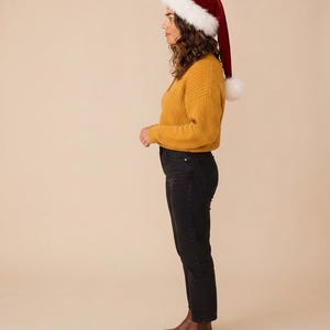 May include: A red velvet Santa hat with a white faux fur trim and pom-pom, a mustard yellow ribbed long-sleeve sweater, black denim jeans, and brown leather ankle boots. The outfit is shown from a side profile view against a neutral background.