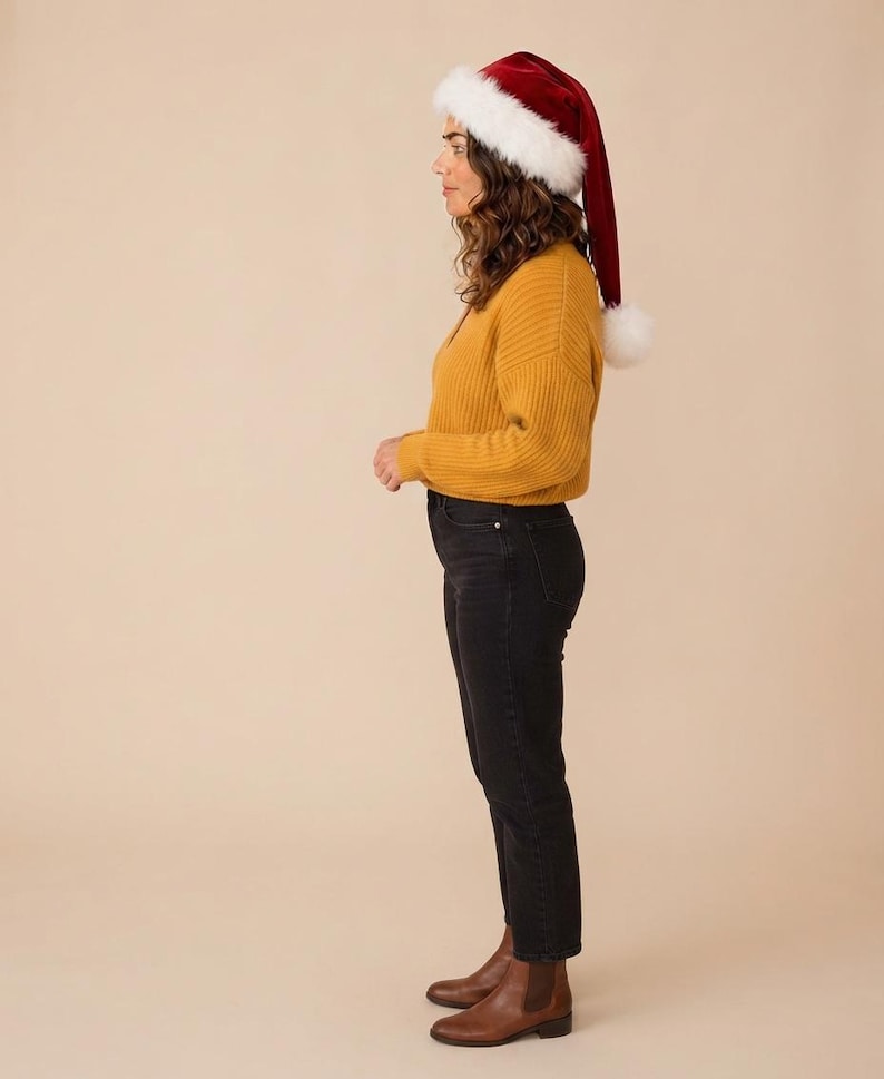 May include: A red velvet Santa hat with a white faux fur trim and pom-pom, a mustard yellow ribbed long-sleeve sweater, black denim jeans, and brown leather ankle boots. The outfit is shown from a side profile view against a neutral background.