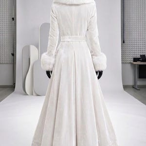 以下が含まれることがあります： 白いベルベット素材の床まで届くロングコート。幅の広い白いフェイクファーの襟と、長袖の袖口にはお揃いのフェイクファーがあしらわれています。ベルト付きのウエスト、プリーツ入りのフレアスカートが特徴で、黒い手袋を着用しています。
