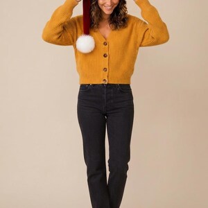 May include: A mustard yellow ribbed knit cardigan with a V-neckline and four brown buttons, paired with black denim jeans and brown leather boots. A red velvet Santa hat with a white faux fur trim and a white pom-pom is worn on the head.