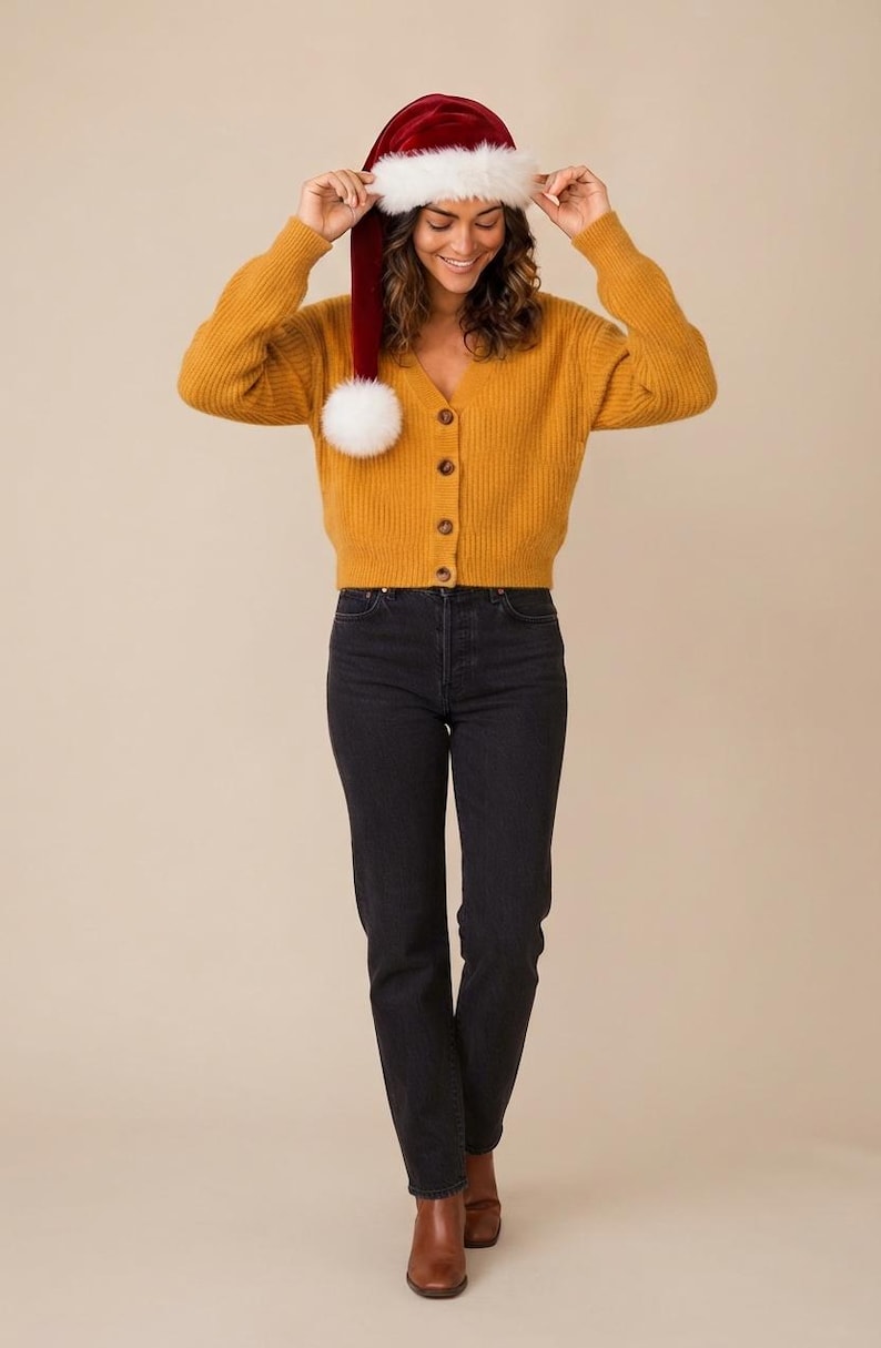 May include: A mustard yellow ribbed knit cardigan with a V-neckline and four brown buttons, paired with black denim jeans and brown leather boots. A red velvet Santa hat with a white faux fur trim and a white pom-pom is worn on the head.