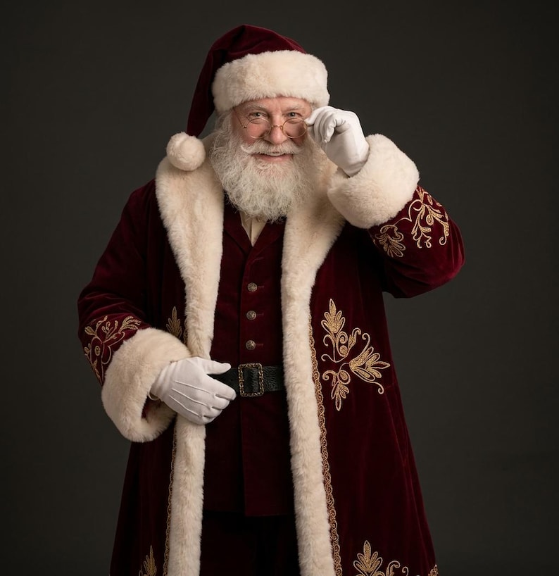 May include: A Santa Claus figure wearing a deep red velvet coat with gold embroidery and thick white faux fur trim. The outfit includes a matching red hat with a white pom-pom, a dark vest, a black belt with a buckle, and white gloves.