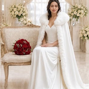 May include: A floor-length white wedding dress with a V-neckline and a matching white cape featuring a thick faux fur collar and trim. The cape drapes over the shoulders and extends to the floor, creating a long train. A bouquet of red roses sits nearby.