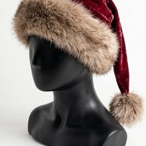 Puede incluir: Un gorro de terciopelo rojo intenso con un borde ancho de piel sint&eacute;tica beige esponjosa en la base y un pomp&oacute;n de piel sint&eacute;tica a juego unido al extremo de la parte superior puntiaguda y ca&iacute;da, sobre una cabeza de maniqu&iacute; negra y lisa.