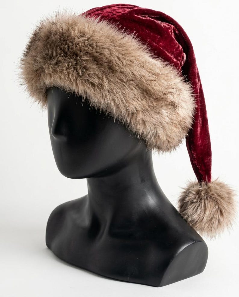 Puede incluir: Un gorro de terciopelo rojo intenso con un borde ancho de piel sint&eacute;tica beige esponjosa en la base y un pomp&oacute;n de piel sint&eacute;tica a juego unido al extremo de la parte superior puntiaguda y ca&iacute;da, sobre una cabeza de maniqu&iacute; negra y lisa.