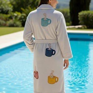May include: A light gray bathrobe featuring embroidered patches of four coffee mugs in teal, dark blue, red, and yellow. Each mug patch includes a swirling steam design rising from the top. The robe has long sleeves, a shawl collar, and a tie belt.
