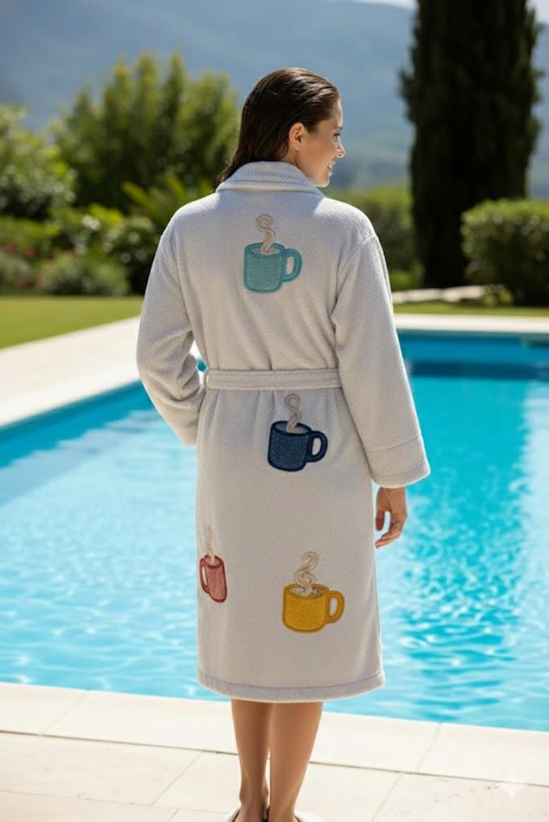 May include: A light gray bathrobe featuring embroidered patches of four coffee mugs in teal, dark blue, red, and yellow. Each mug patch includes a swirling steam design rising from the top. The robe has long sleeves, a shawl collar, and a tie belt.